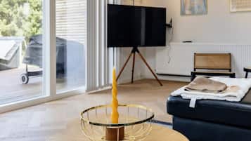 A modern living room with a television on a tripod stand, a round glass table with a candle, a black sofa, and a window with blinds.