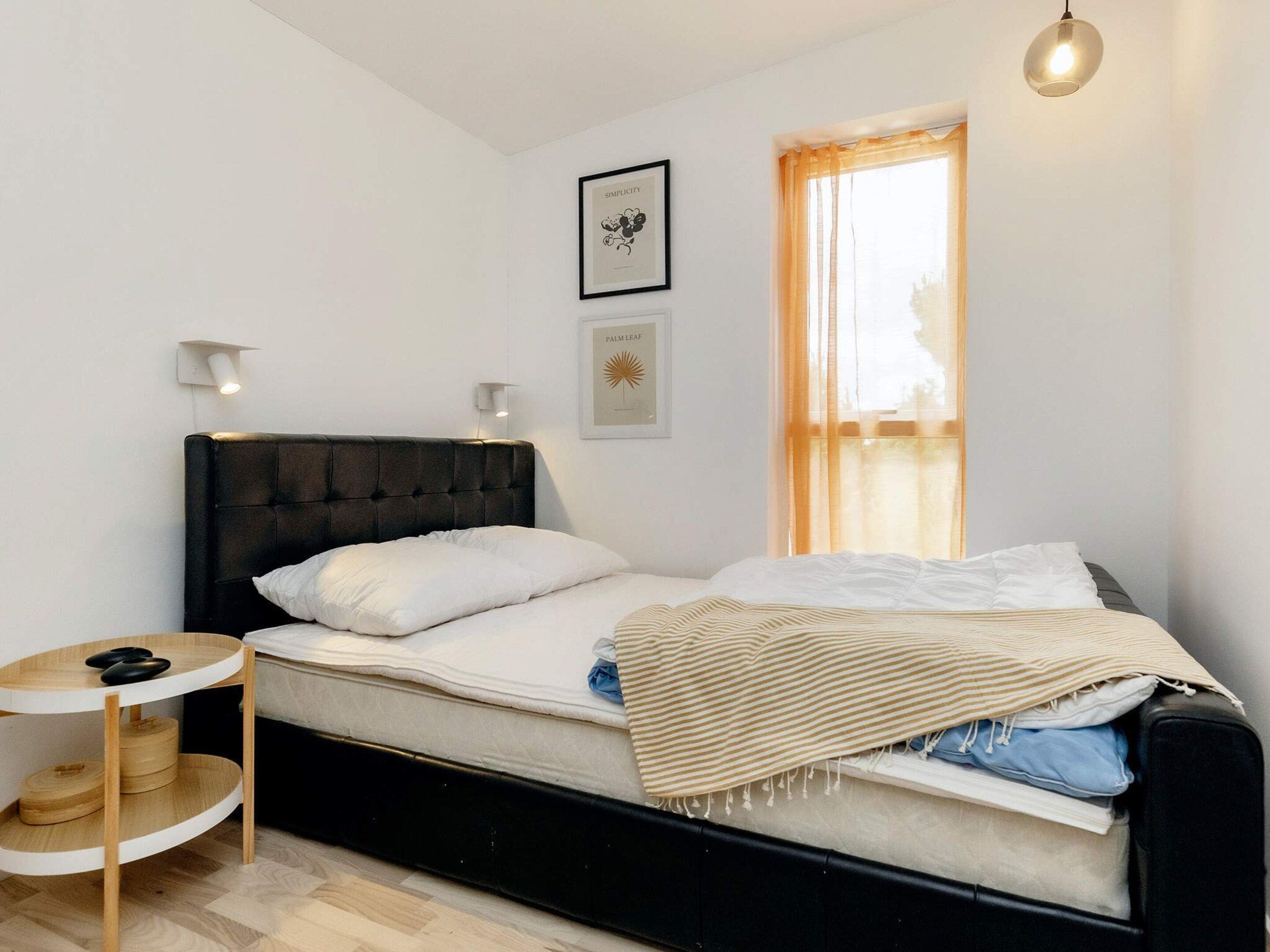 A single bed with a black headboard, white bedding, a small round wooden side table, and a wall-mounted light fixture.