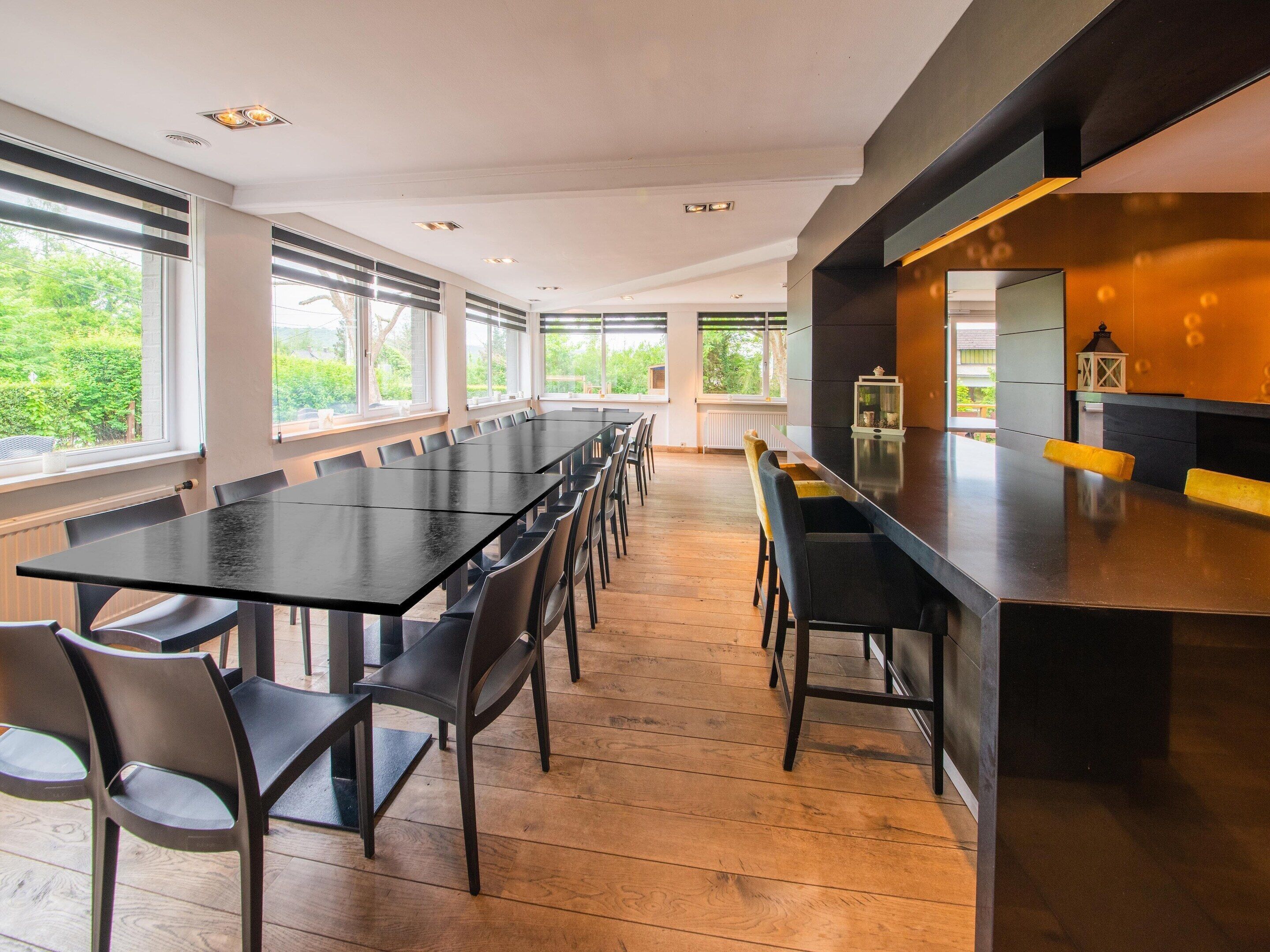 Un espace repas moderne avec une longue table et des chaises, de grandes fenêtres et un coin cuisine doté d’un bar.