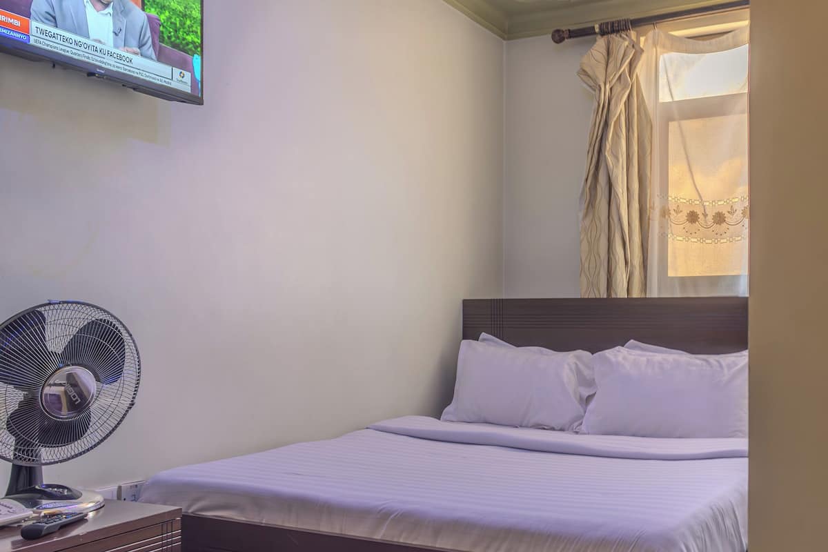 Un dormitorio con cama, una mesita de noche con un ventilador, un televisor montado en la pared y una ventana con cortinas.