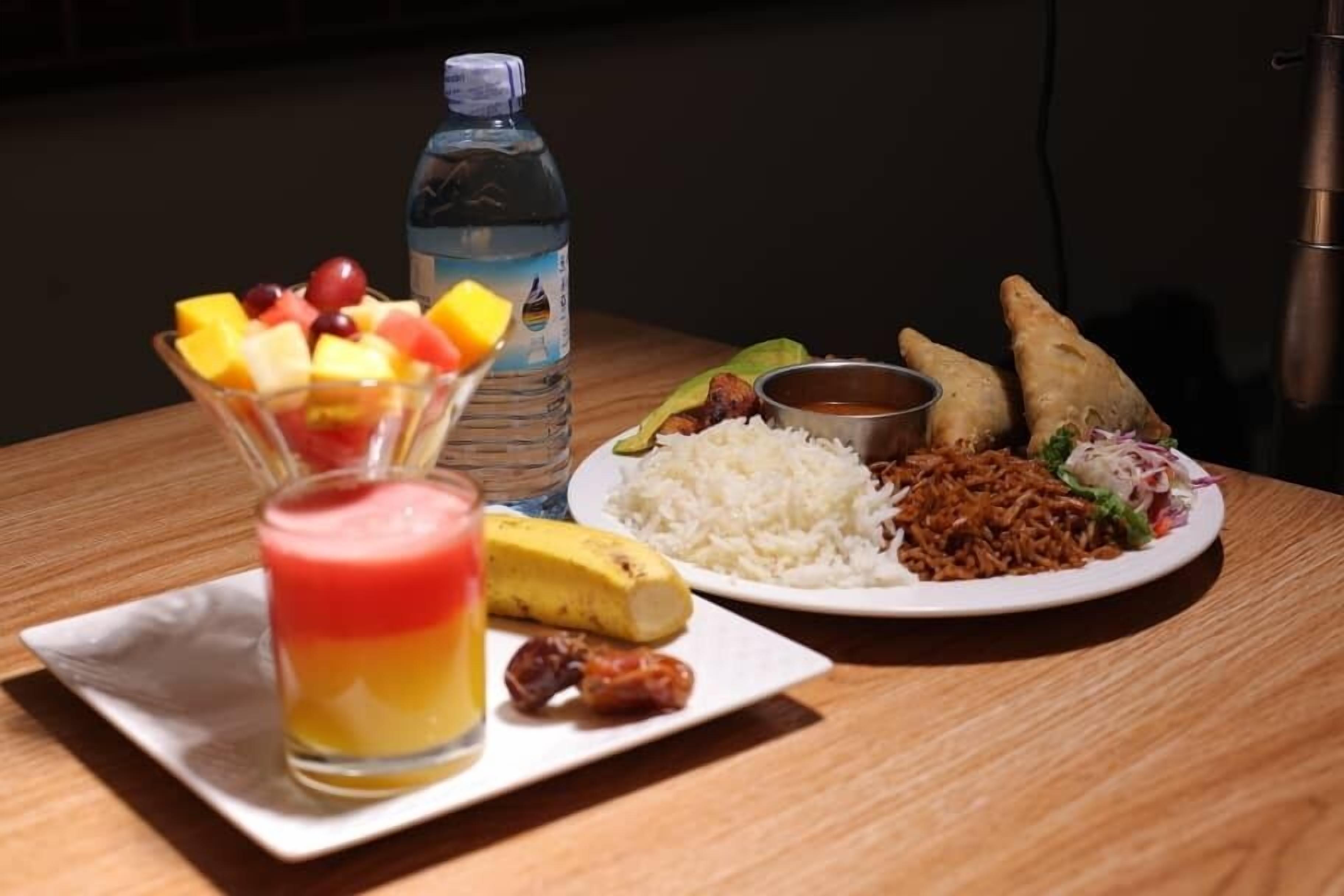 A meal with rice, meat, and vegetables, a banana, a fruit salad, and a bottle of water on a wooden table.