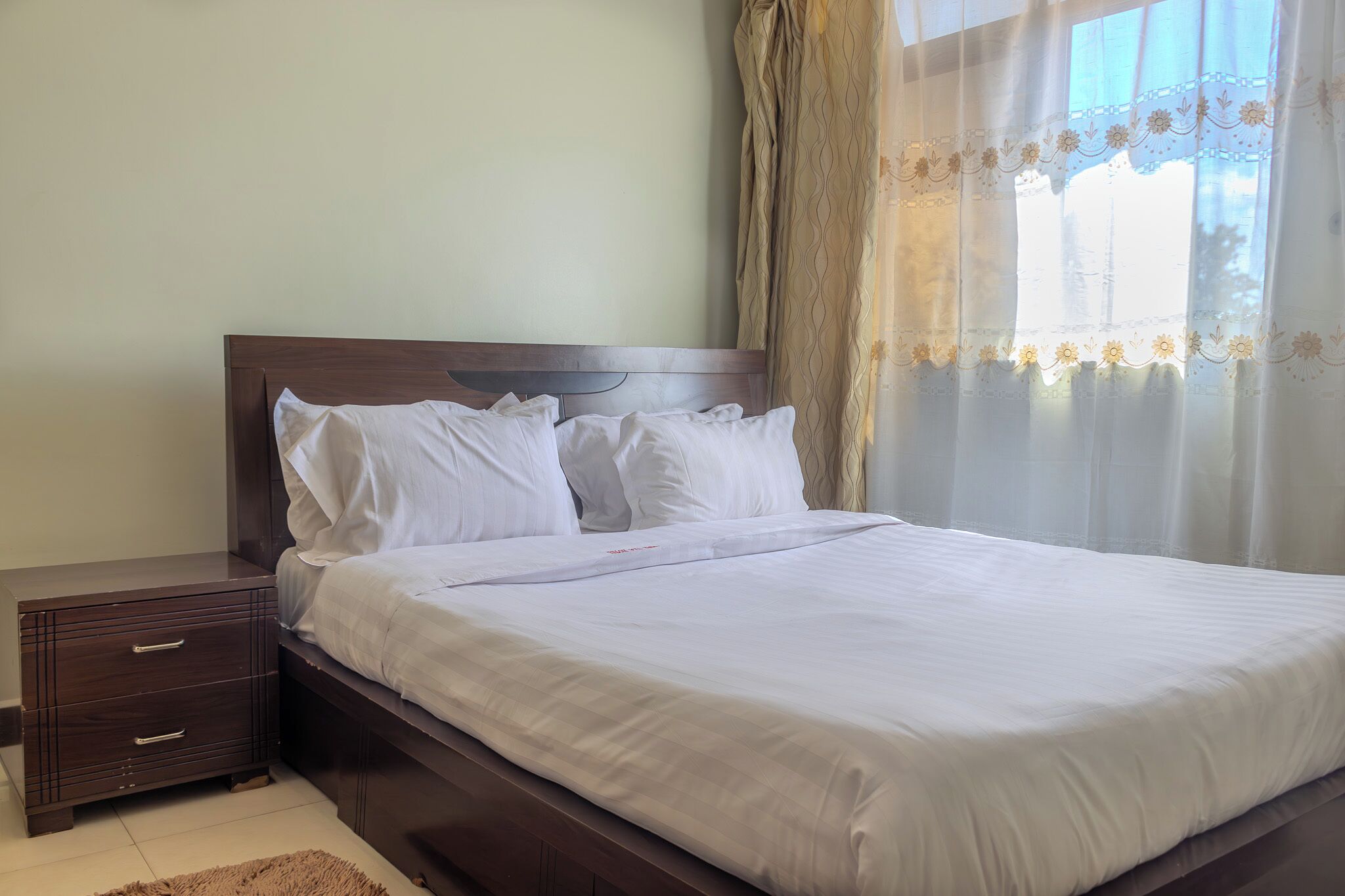 A neatly made bed with white linens in a room with a wooden headboard and a nightstand with drawers.