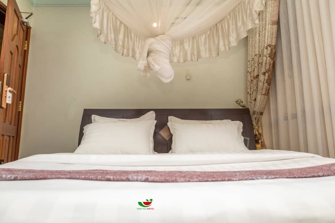 A neatly made bed with white linens and a pink accent, a dark headboard, and a canopy overhead.
