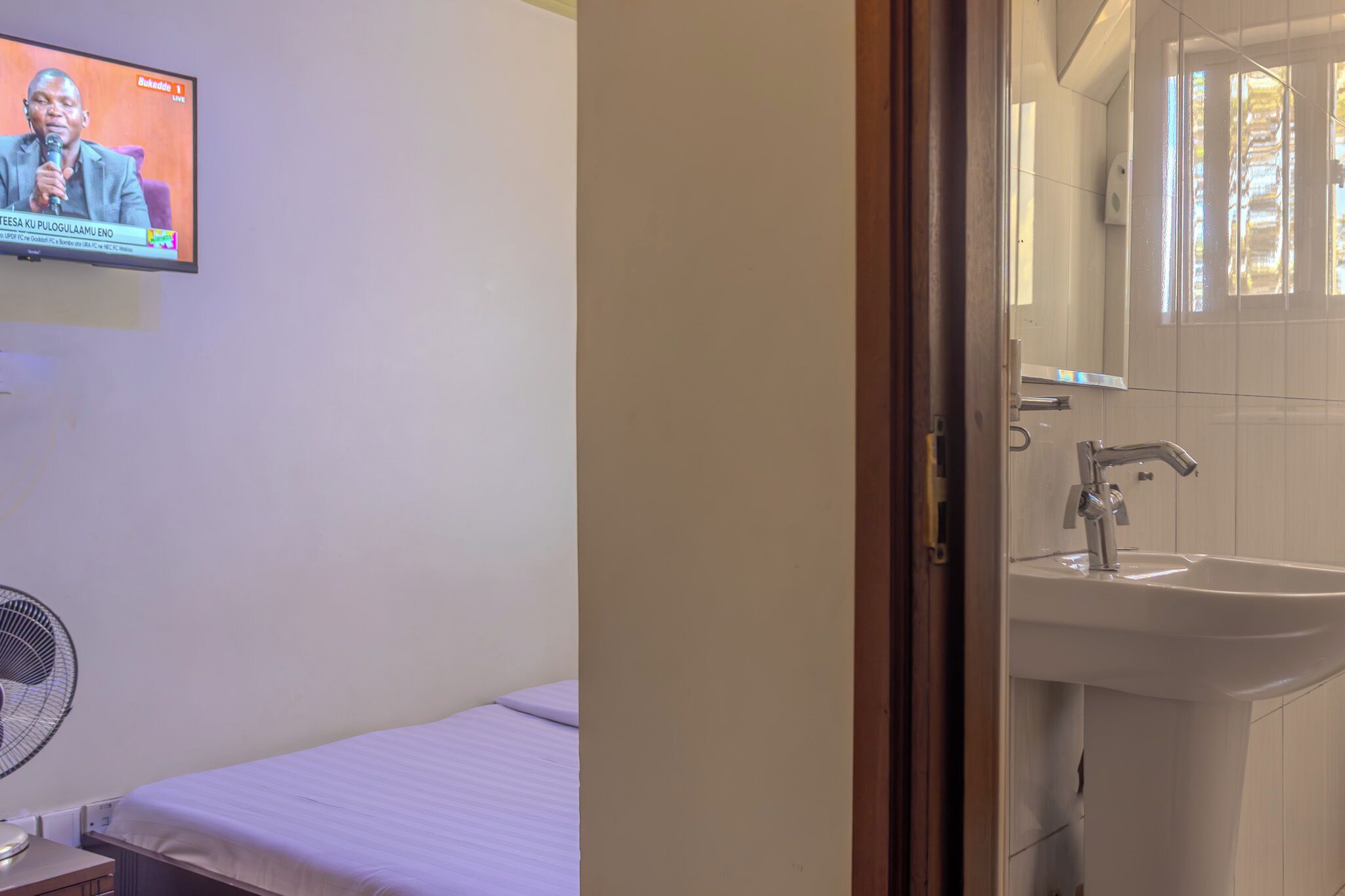 A hotel room with a bed, a wall-mounted TV, a fan, and a bathroom with a sink and mirror.