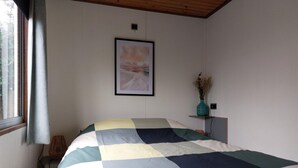Un dormitori amb un llit gran, un quadre al mur, una petita estanteria amb un vas i una finestra amb cortines.