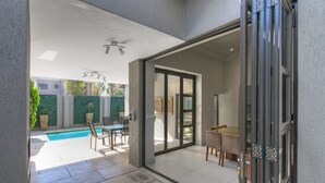Ein offener Wohnbereich mit Glastür zu einer Terrasse mit Pool, Gartenmöbeln und Blick auf ein Nachbarhaus.