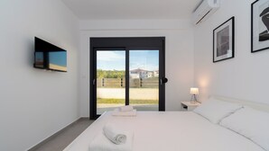 A modern hotel room with a bed, a TV mounted on the wall, a bedside table with a lamp, and a large window with a view of buildings and greenery. - SithoniaRS Ground Floor Gem in Nikiti (Nikiti)
