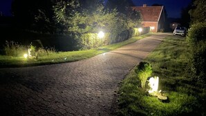 A paved driveway leading to a house with a red roof, surrounded by greenery and illuminated by street lamps. - Shell Modern Retreat (Dornum)