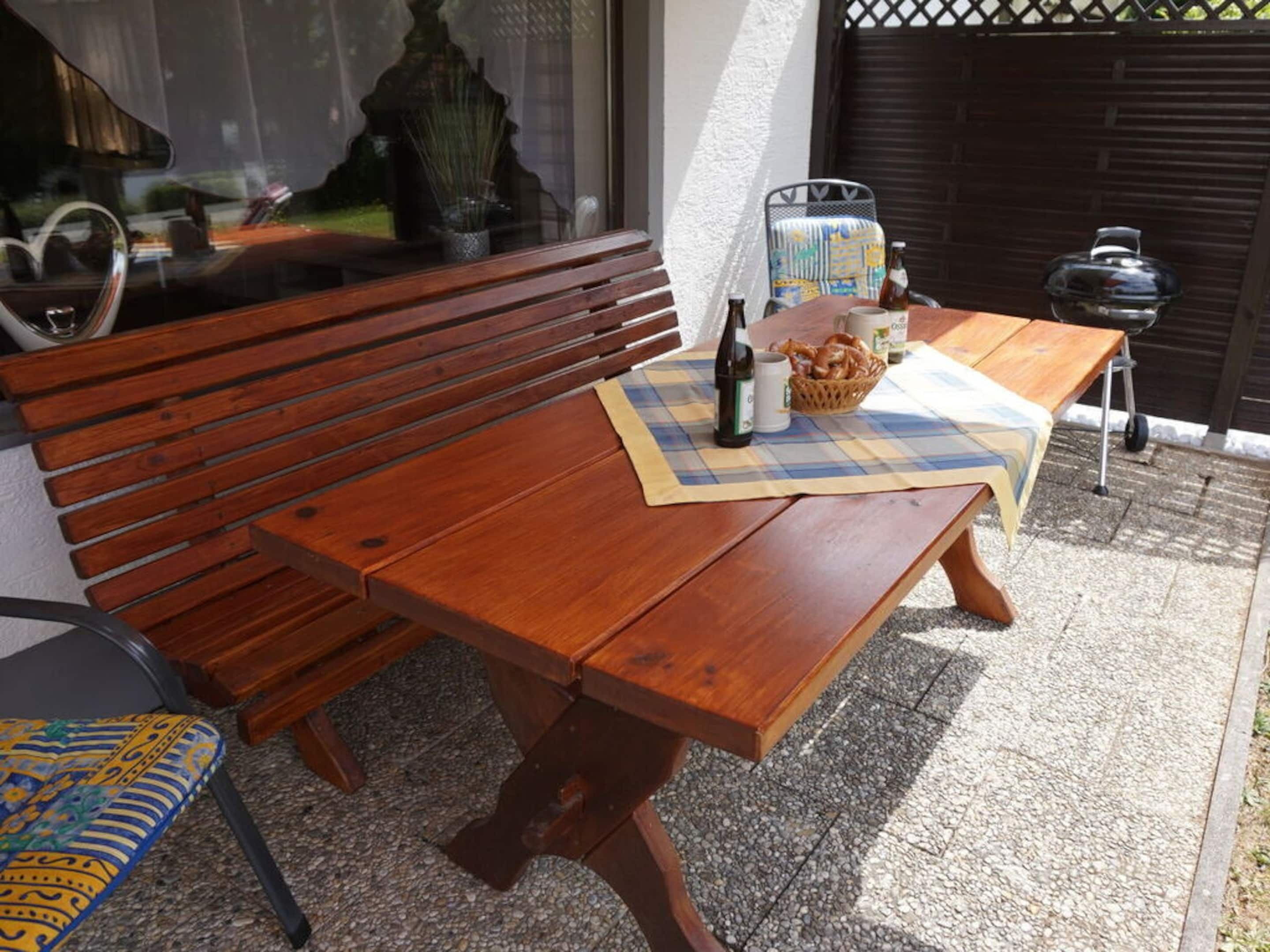 A wooden bench and table set on a patio with a wicker basket, bottles, and a placemat.