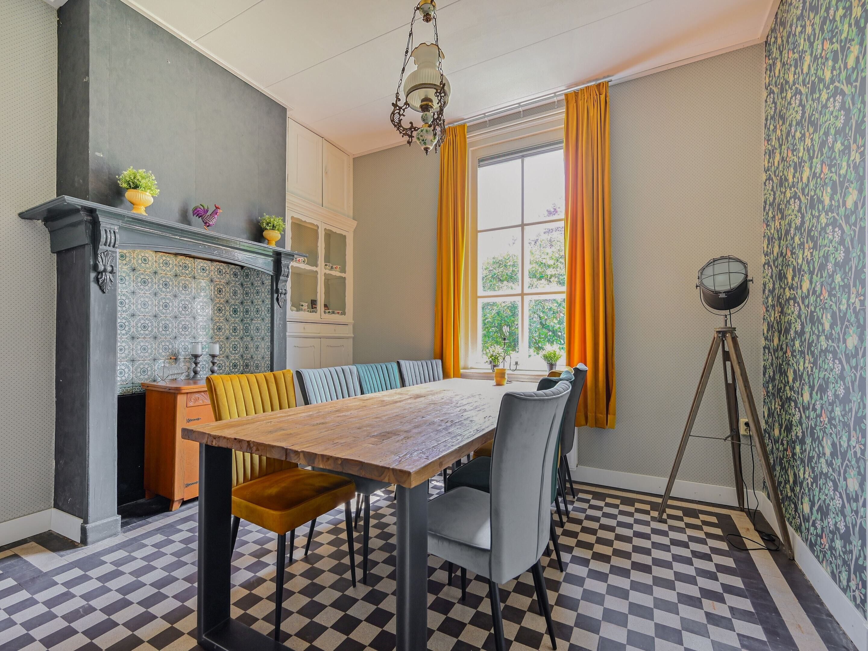 A dining area with a checkered floor, a wooden table, and chairs. There is a window with curtains, a chandelier, and a cabinet with glass doors.