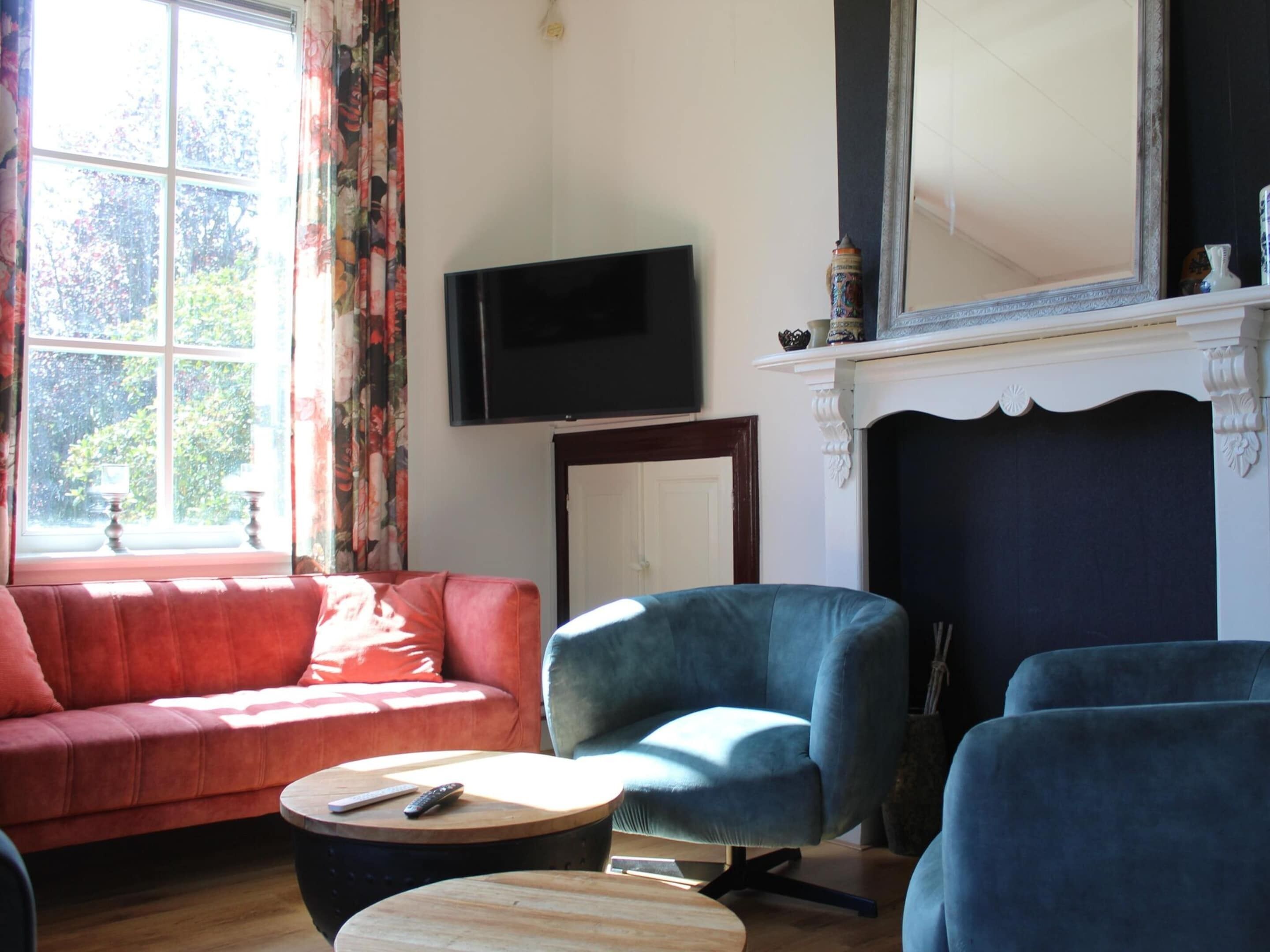 Un salon avec un canapé rouge, un fauteuil bleu et une table basse en bois. Un téléviseur à écran plat est fixé au mur, au-dessus d’une cheminée.