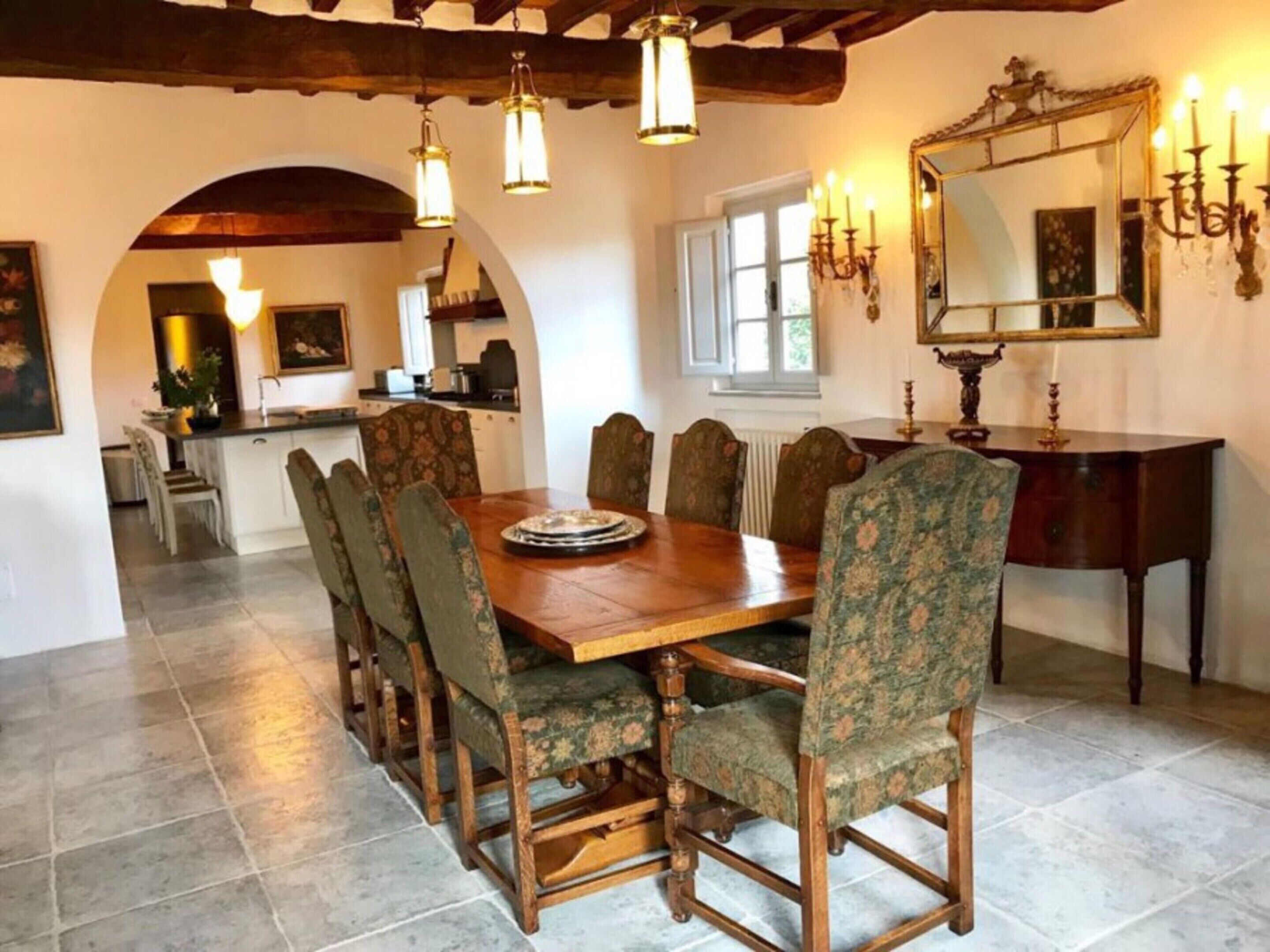 Un comedor con una mesa de madera, sillas tapizadas con motivos florales, una araña y un espejo en la pared.