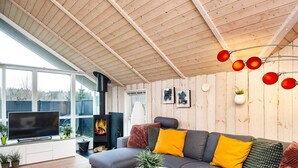 Een moderne woonkamer met een grote grijze bank, een witte salontafel en een televisie op een standaard. Er is een haard met een zwart rookkanaal, en de kamer heeft houten wanden en een hoge plafond.