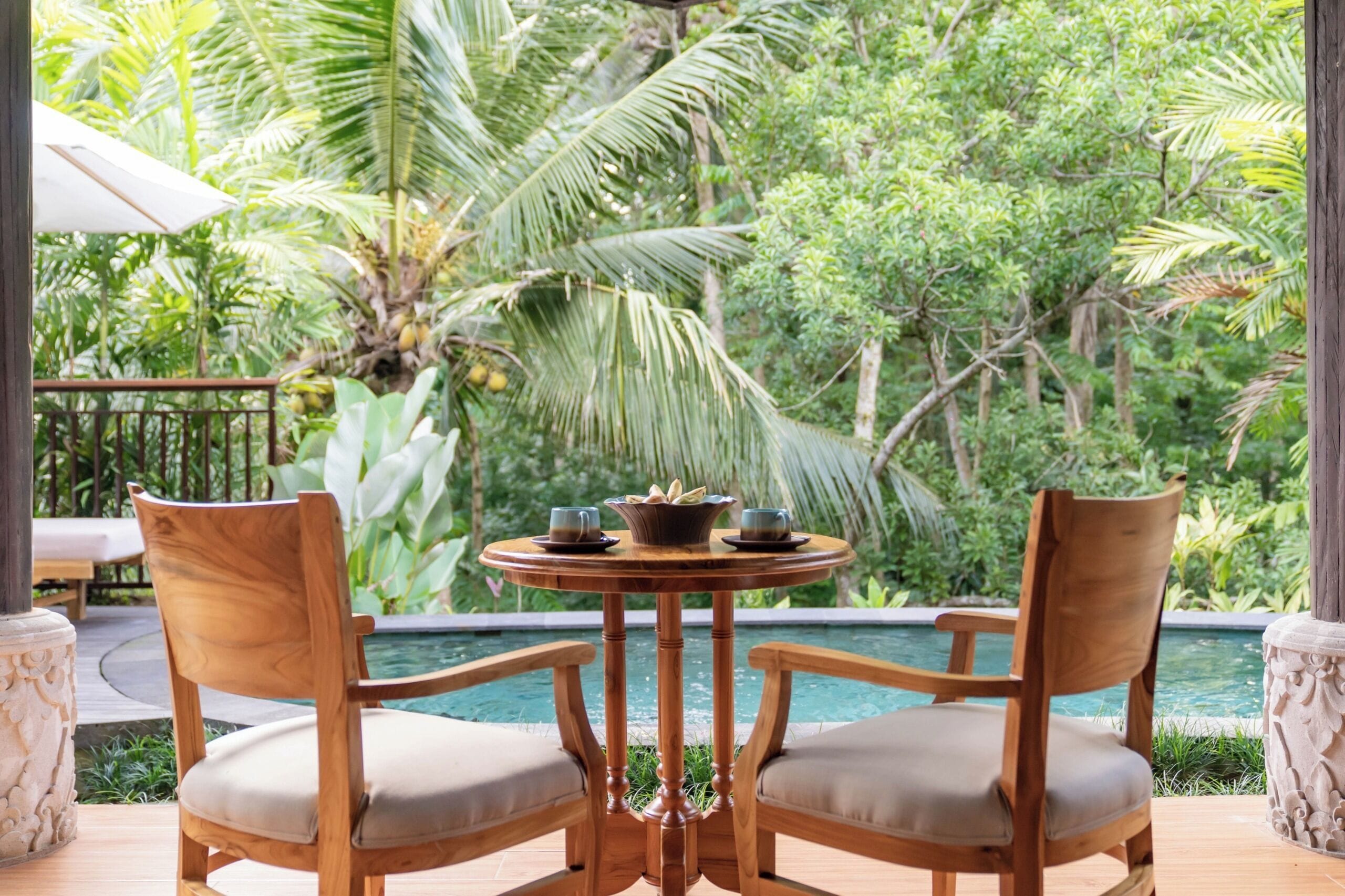 A wooden table with two chairs and a small table centerpiece, overlooking a pool with lush greenery in the background.