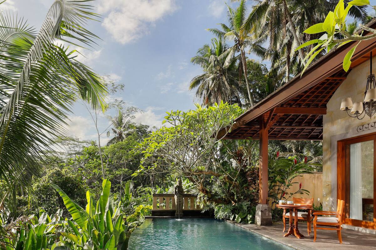A poolside area with a lounge chair, a wooden table with chairs, and a view of the pool surrounded by tropical vegetation.