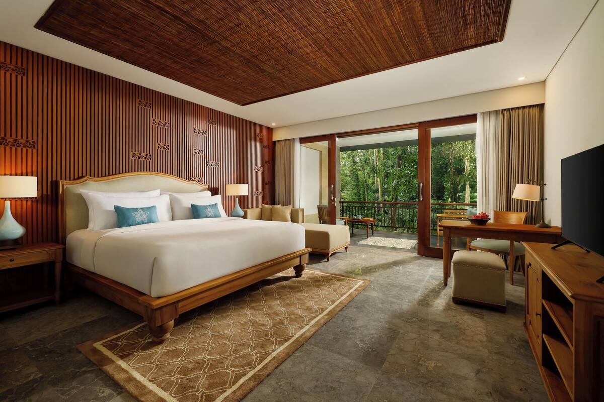 A spacious bedroom with a large bed, a desk, and a balcony with a view of greenery.