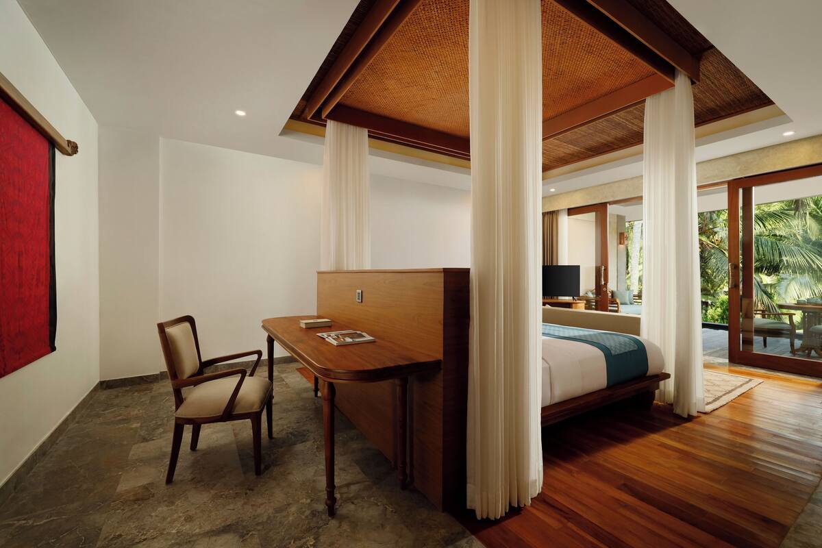 A bedroom with a bed, a desk, and a chair. There is a window with a view of palm trees.
