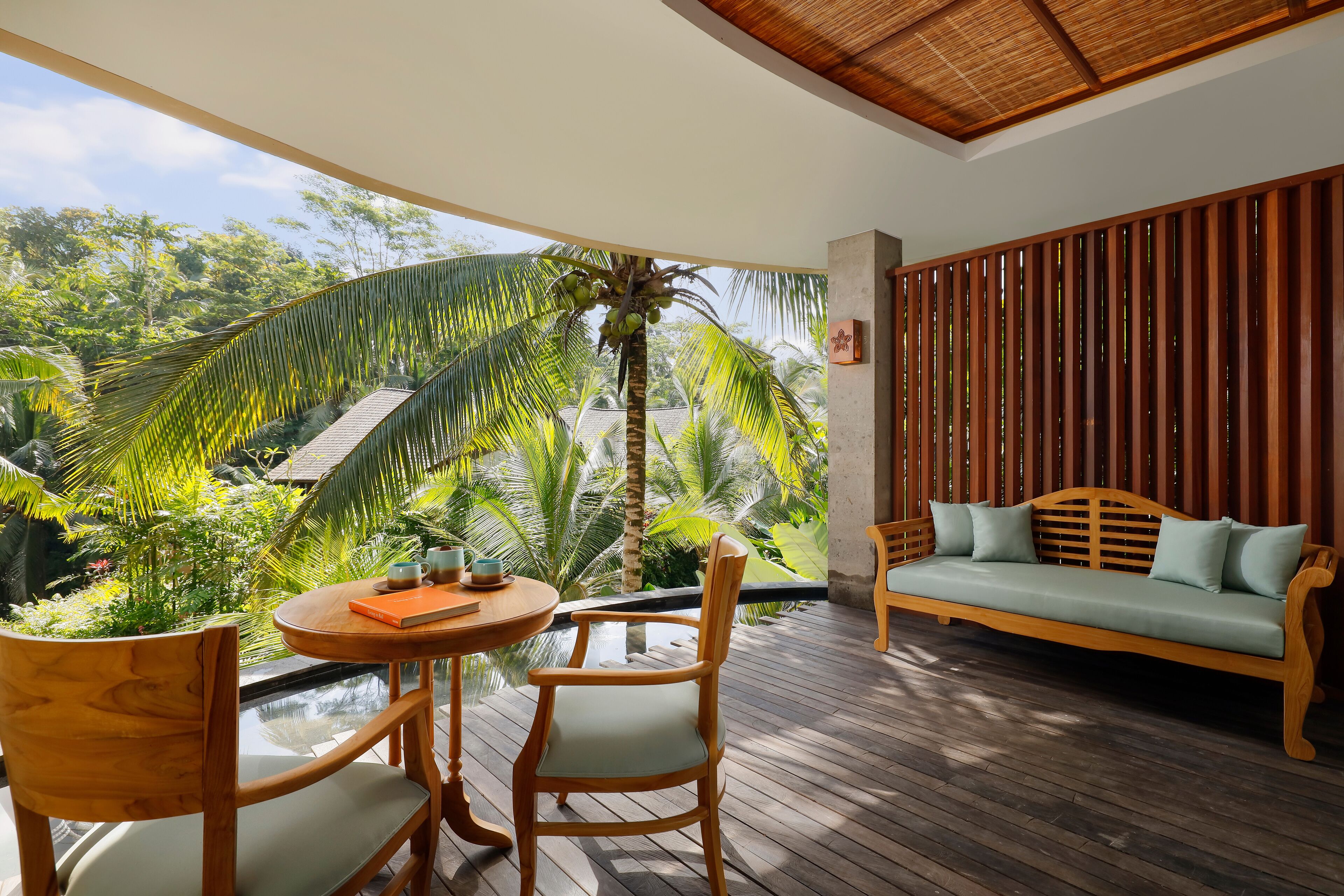 A wooden deck with a sofa and chairs, overlooking a lush garden with palm trees.