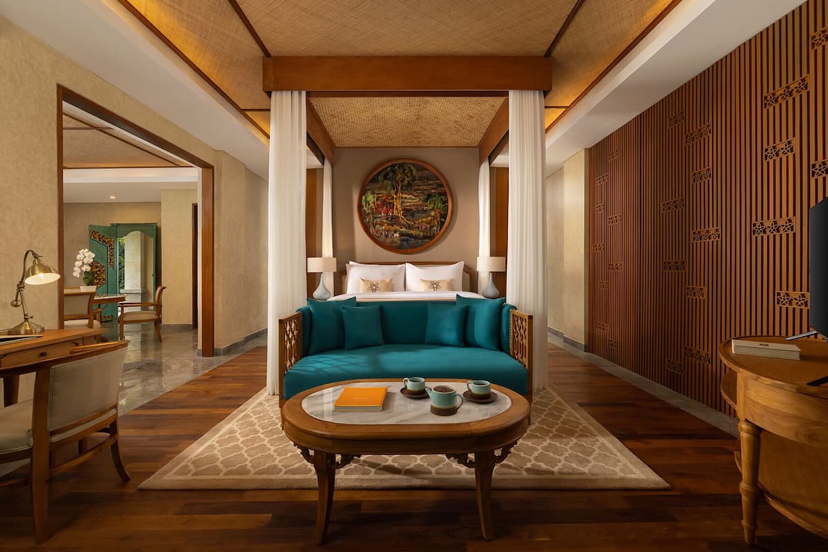 A hotel room with a teal sofa, a coffee table with a book and teacups, a painting on the wall, and a wooden panel wall.