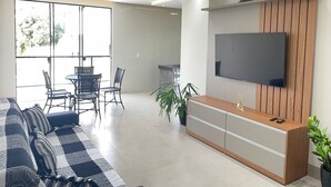 A modern living room with a sofa, a dining table with chairs, a TV mounted on a wooden panel wall, and a potted plant. - Desfrute de Momentos Inesquecíveis em Caldas Novas! Casa Aconchegante 3 Suites (Portal das Águas Quentes Caldas Novas)