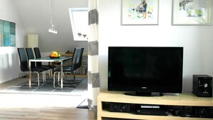 A modern living room with a flat-screen TV on a wooden stand, a dining area with a table and chairs, and artwork on the walls.