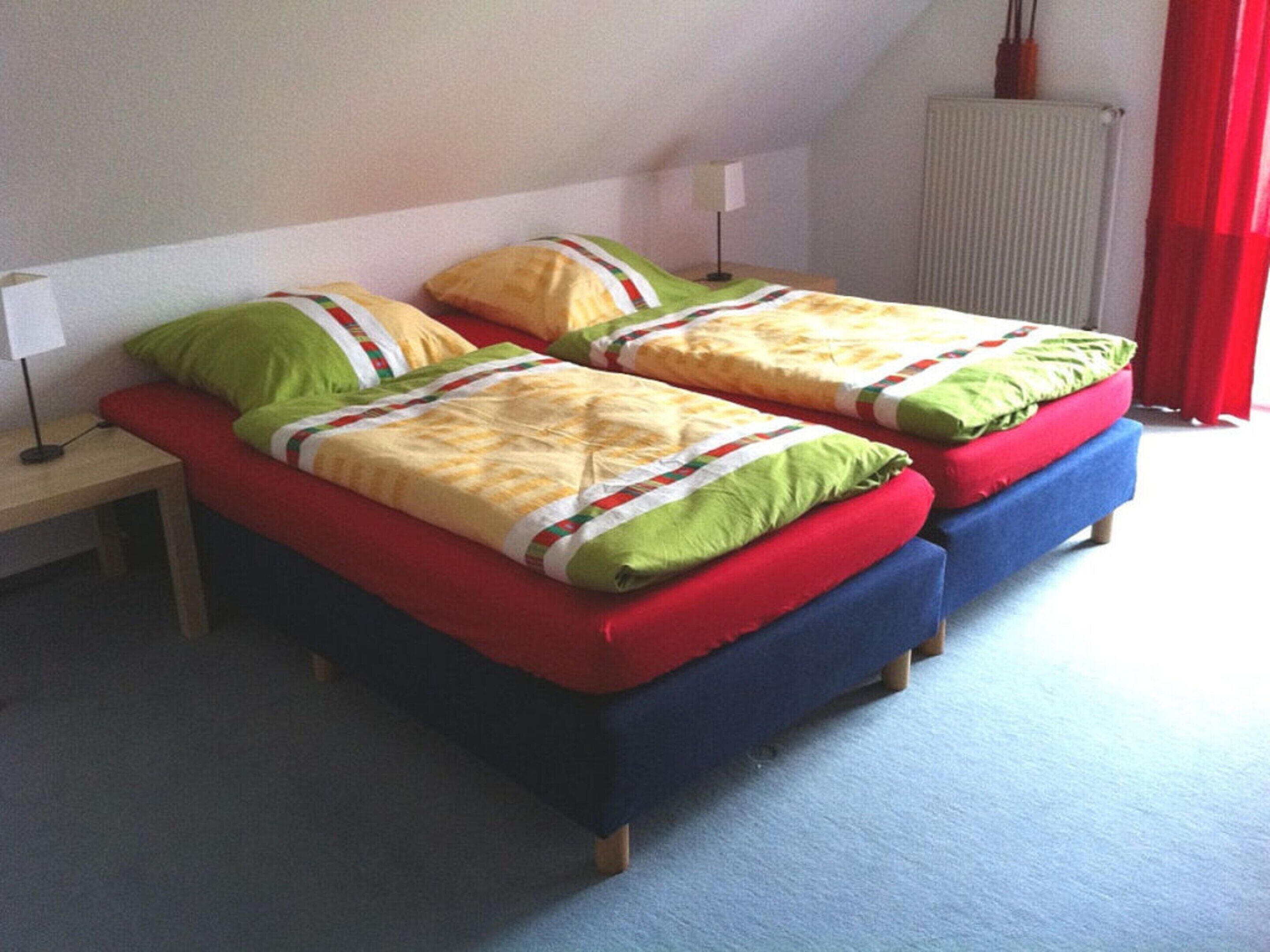 A room with two single beds, each with a red mattress and green pillows. There is a small table with a lamp beside one bed, and a radiator on the wall.