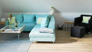 A modern living room with a teal sofa, a glass coffee table, a black armchair, and a small white dog on the sofa.