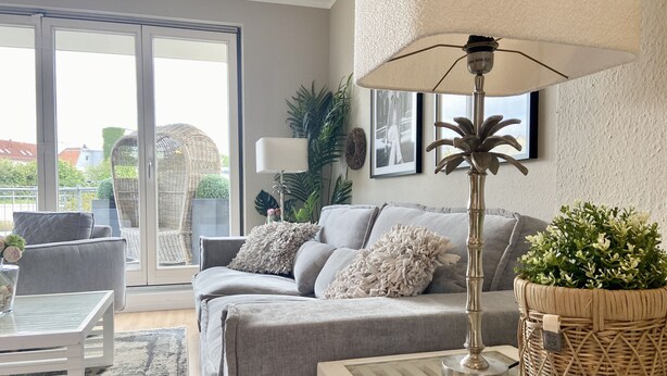 Una sala de estar moderna con un sofá gris, una mesa de centro blanca y una mesita auxiliar de mimbre con una planta.