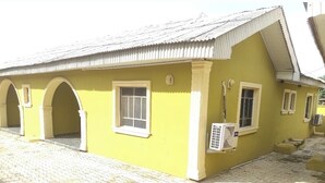 Un bâtiment jaune avec un toit en tuiles, une porte d’entrée centrale et une fenêtre équipée d’un appareil de climatisation.