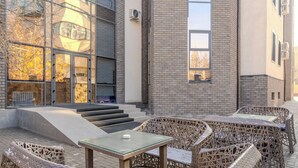 An outdoor seating area with wicker furniture and a glass table, adjacent to a building with large windows and a brick facade.