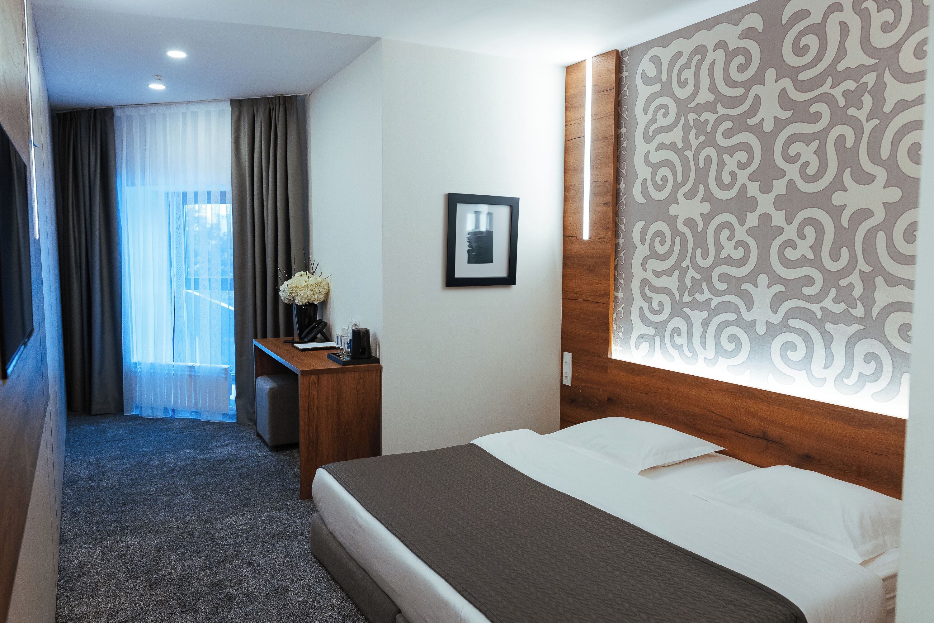 A hotel room with a bed, a desk with a chair, a picture on the wall, and a patterned wall panel.