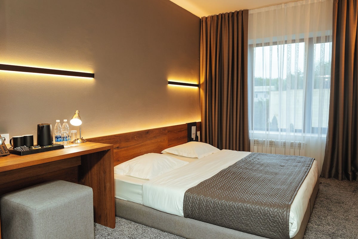 A hotel room with a bed, bedside table, lamp, and a view of the outdoors.