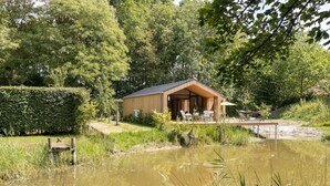 A wooden cabin with a covered patio, surrounded by lush greenery and a pond. - Nice Holiday Home in Kortgene With Garden (Kortgene)