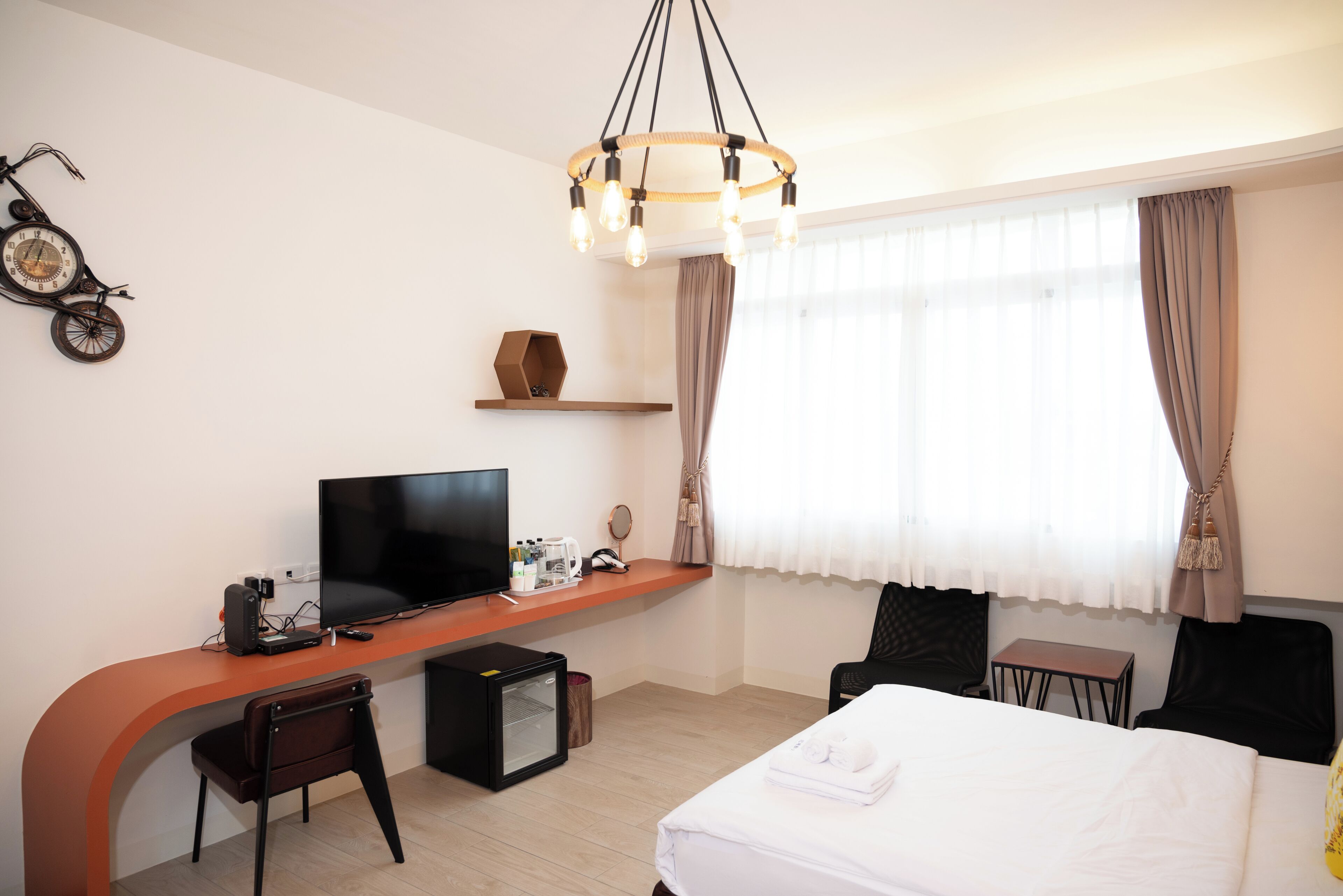 A modern hotel room with a curved desk, a flat-screen TV, a bed with white linens, and a large window with curtains.