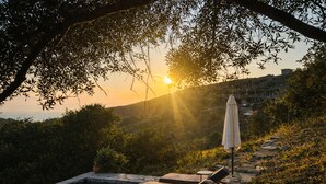 A stone pool with lounge chairs and an umbrella, surrounded by greenery and overlooking a hillside. - Villa Kotseli: Traditional Stone House with Pool & Sunset Sea Views (Andros)