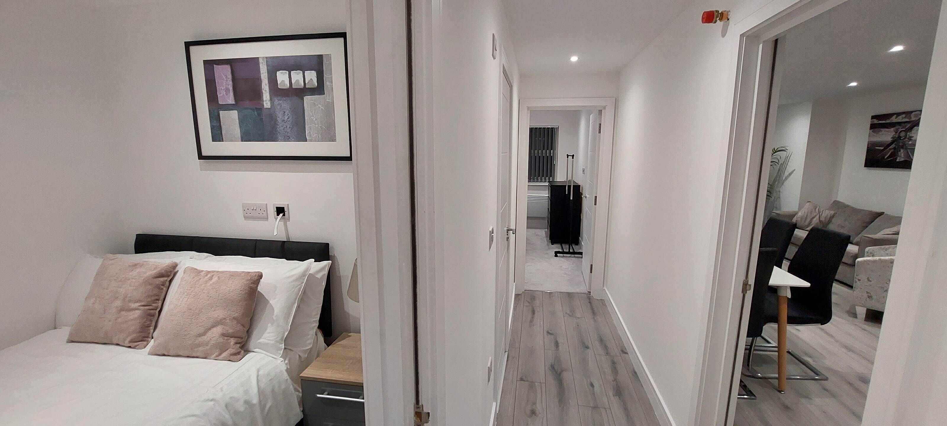 Et hotelværelse med seng, natbord, et billede i ramme på væggen og en gang, der fører til en opholdsstue med sofa og bord.