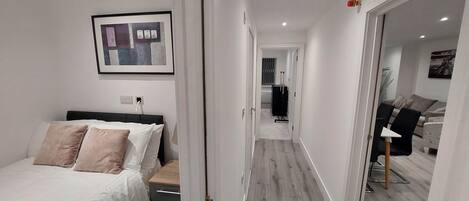 Et hotelværelse med seng, natbord, et billede i ramme på væggen og en gang, der fører til en opholdsstue med sofa og bord.