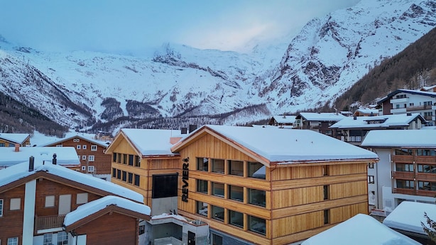A wooden chalet-style building with multiple windows and a snow-covered roof, surrounded by other buildings and snow-covered mountains in the background. - Revier Mountain Lodge Saas-Fee (Saas-Fee)