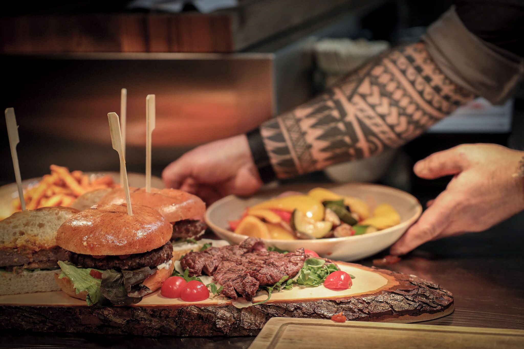 A wooden cutting board with a burger, steak, and a bowl of mixed vegetables.