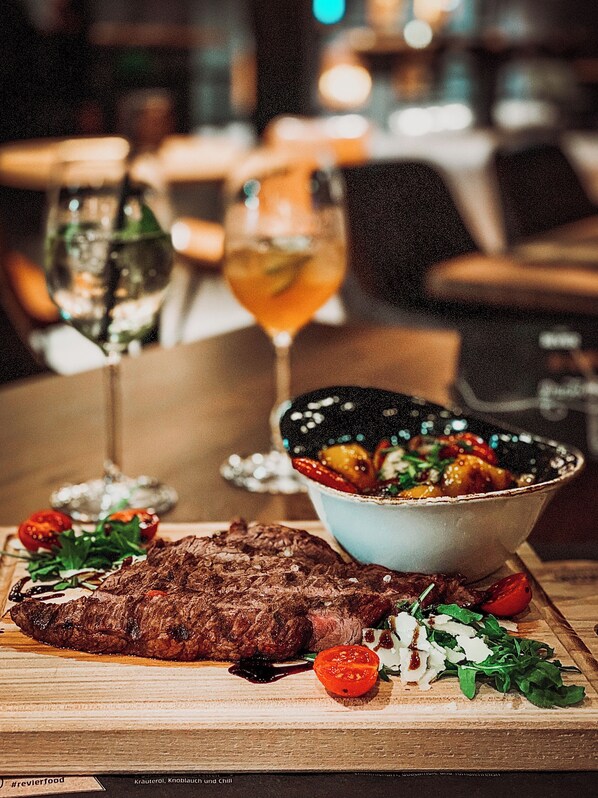 A dining table with a steak, vegetables, and a bowl of salad. There are two glasses of a beverage and a bottle on the table. - Revier Mountain Lodge Saas-Fee (Saas-Fee)