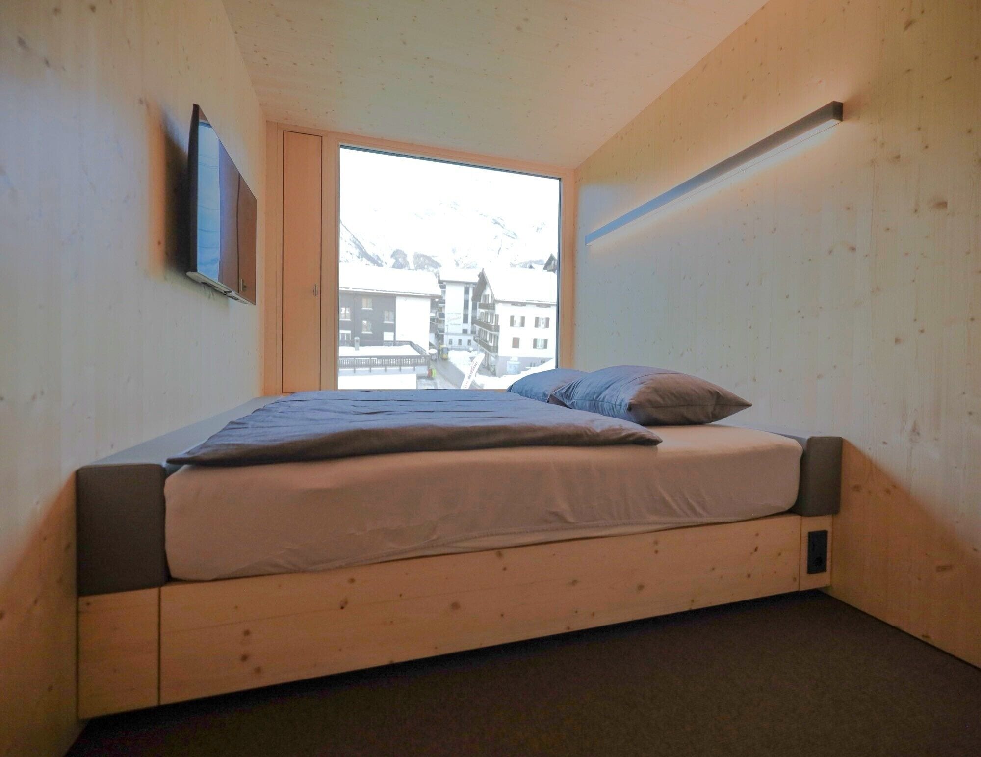A small, wooden-framed room with a single bed, a window with a view of buildings, and a mounted TV.