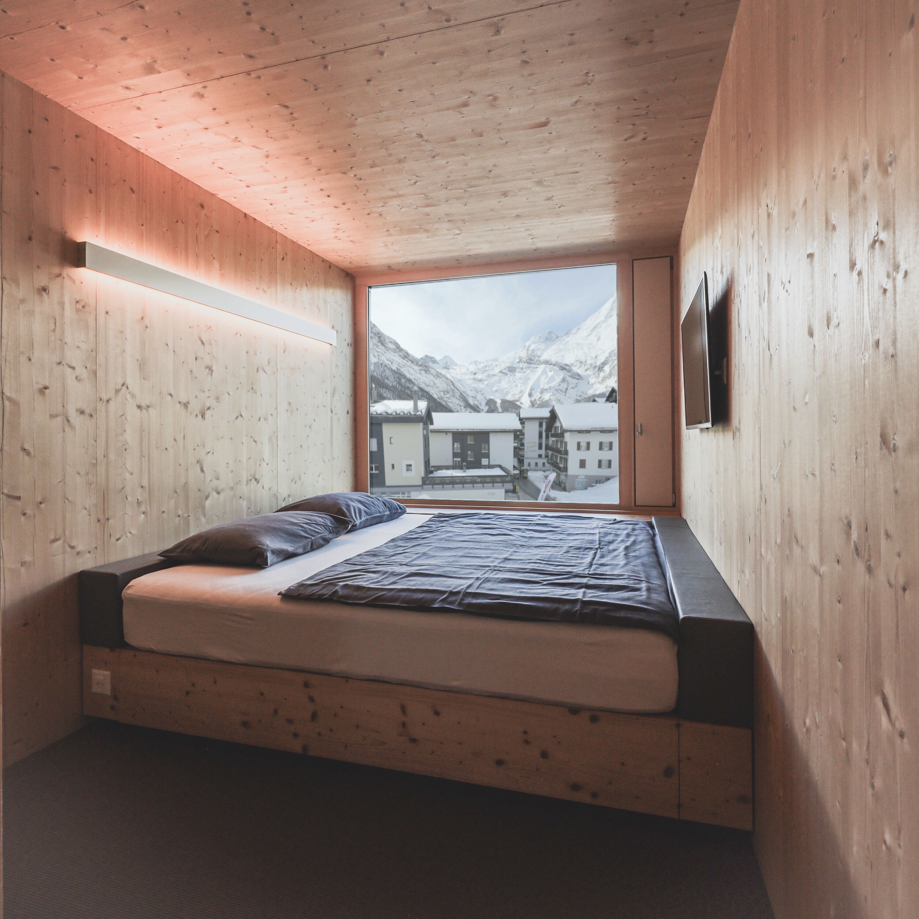 Une chambre avec un plafond en bois, un lit avec un matelas et des oreillers, et une vue sur une montagne et des bâtiments, visible par une fenêtre.