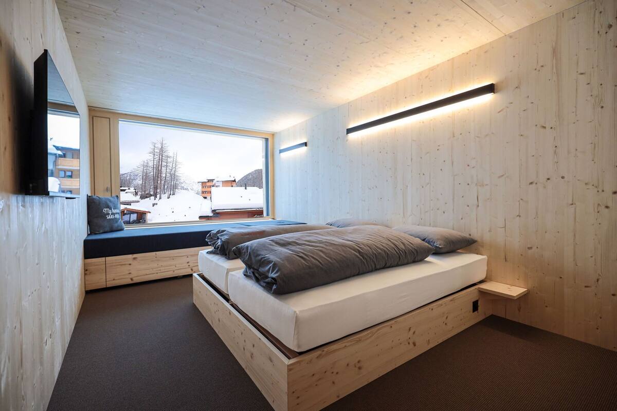 Een moderne slaapkamer met houten vloer, een groot raam met uitzicht op een besneeerd landschap, een bed met grijze beddengoed en een wandgemonteerde televisie.