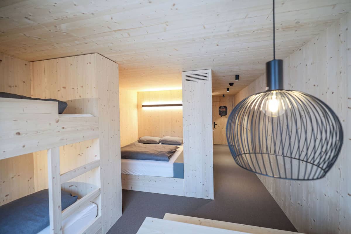 Een kleine, houten kamer met stapelbedden, een enkele bed en een hangende lamp, plus een zichtbare ventilatieopening.