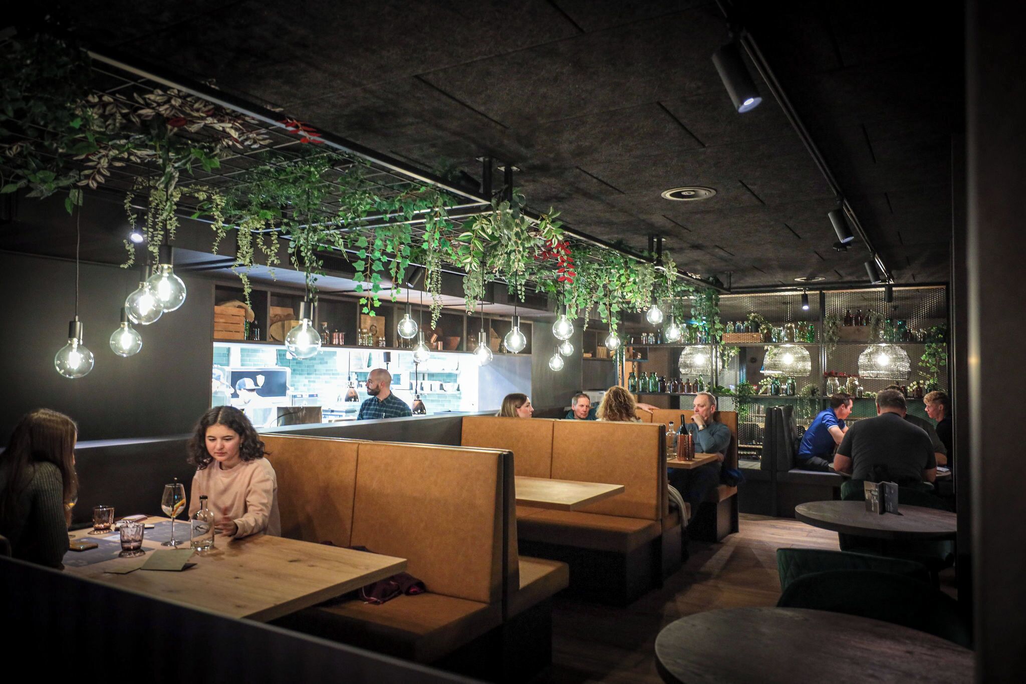 A restaurant with booths and tables, hanging plants, and pendant lighting.