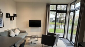 A modern living room with a grey sofa, a round coffee table, and a black armchair. A flat-screen TV is mounted on the wall, and there is a large window with a view of a garden and a body of water. - 55 by the Water (Olburgen)