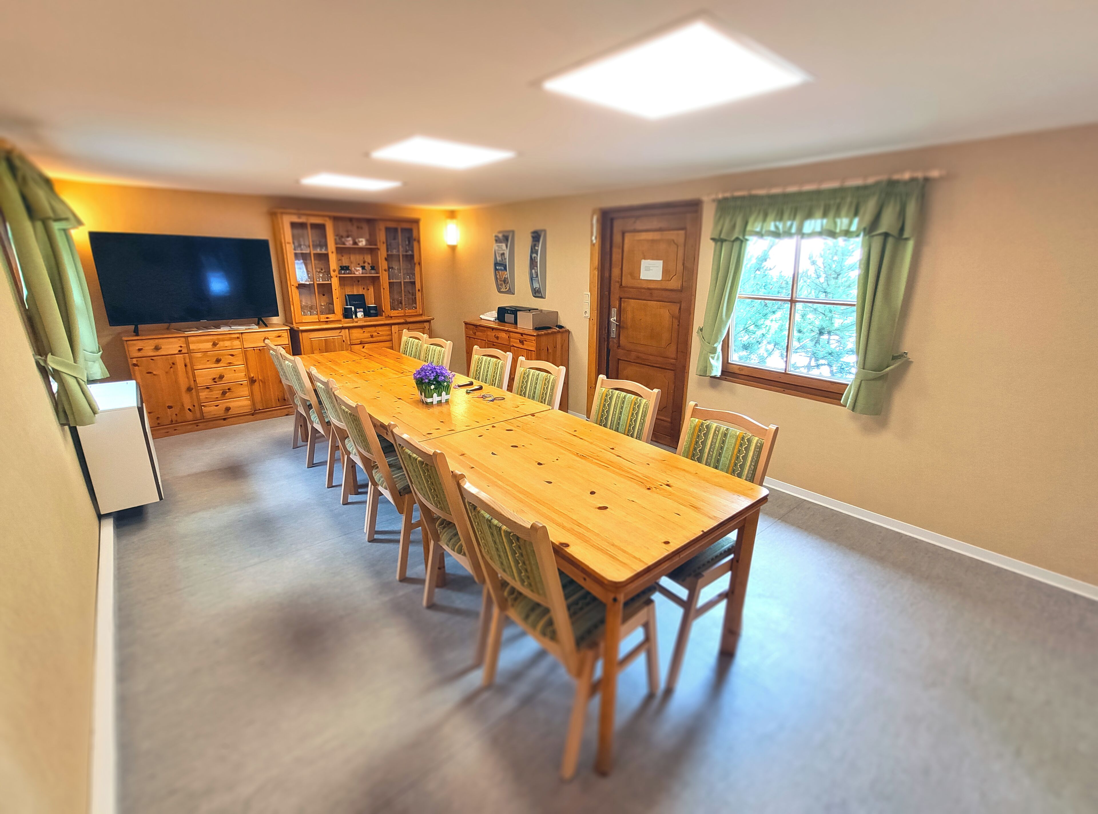 A dining area with a long wooden table, chairs, a television, wooden cabinets, and a window with curtains.