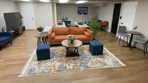 A spacious room with a sofa, two ottomans, a coffee table, and a patterned rug. There are two small tables with plants, a television, and a door in the background. - Conference Room Rental no sleeping arrangements  (Cromwell)