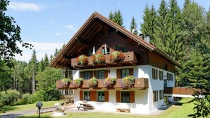 Un chalet de madera de dos pisos con balcones decorados con macetas de flores, un patio con bancos y un paisaje verde exuberante.
