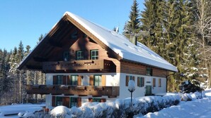 Una cabaña de troncos de dos pisos con techo pronunciado, rodeada de árboles cubiertos de nieve y cielo despejado.